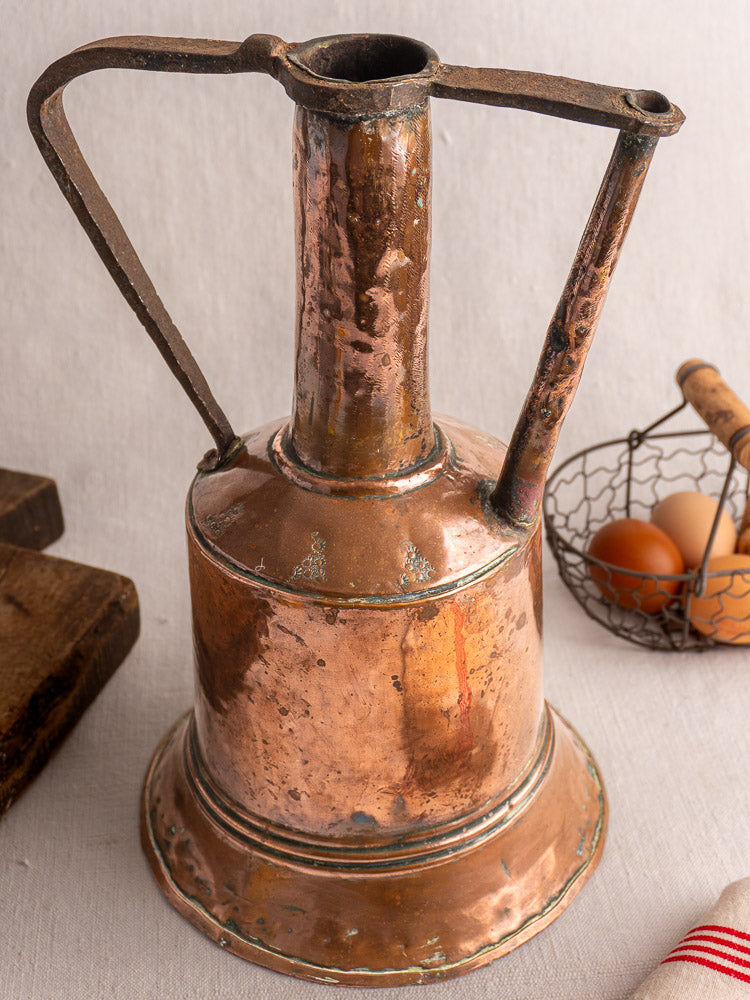 Antique French copper water jug with iron handles and hand-hammered details on a light cloth.