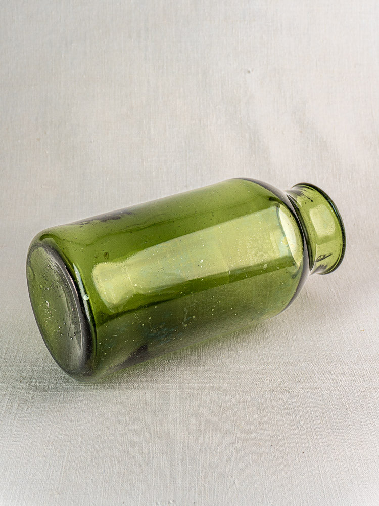 Antique French hand blown green glass cornichon jar with wide base and narrow neck on light gray textured surface.
