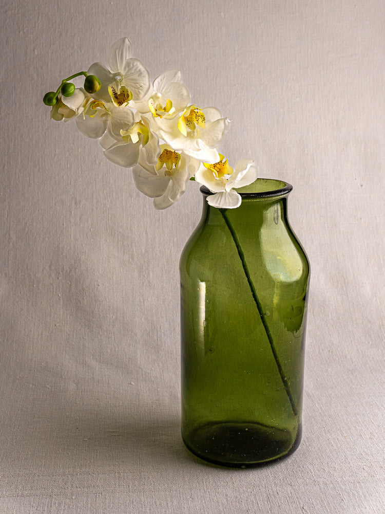 Antique French green glass Cornichon Jar (late 18th century) with white orchid, on light gray fabric background.