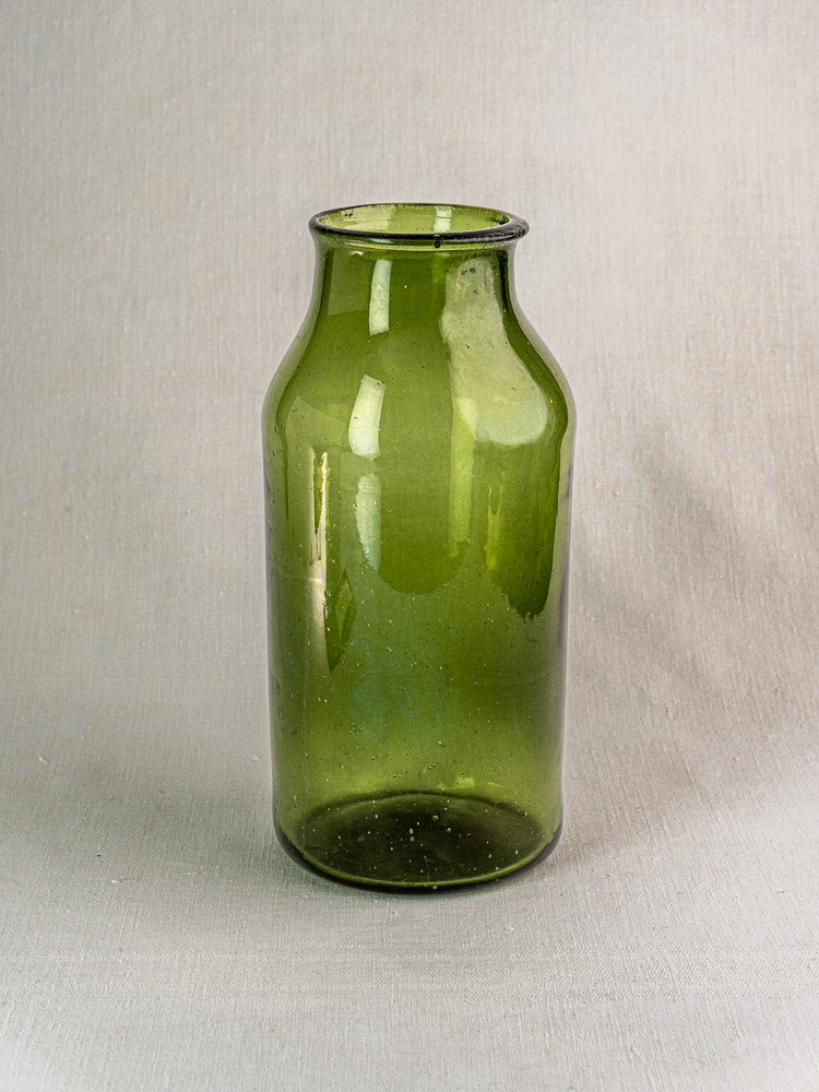 Antique French cornichon jar in hand blown green glass, tall and cylindrical with narrow neck on light textured surface.