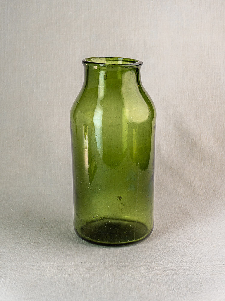 Antique French cornichon jar, hand blown green glass with tall neck and wide body on light gray textured surface.