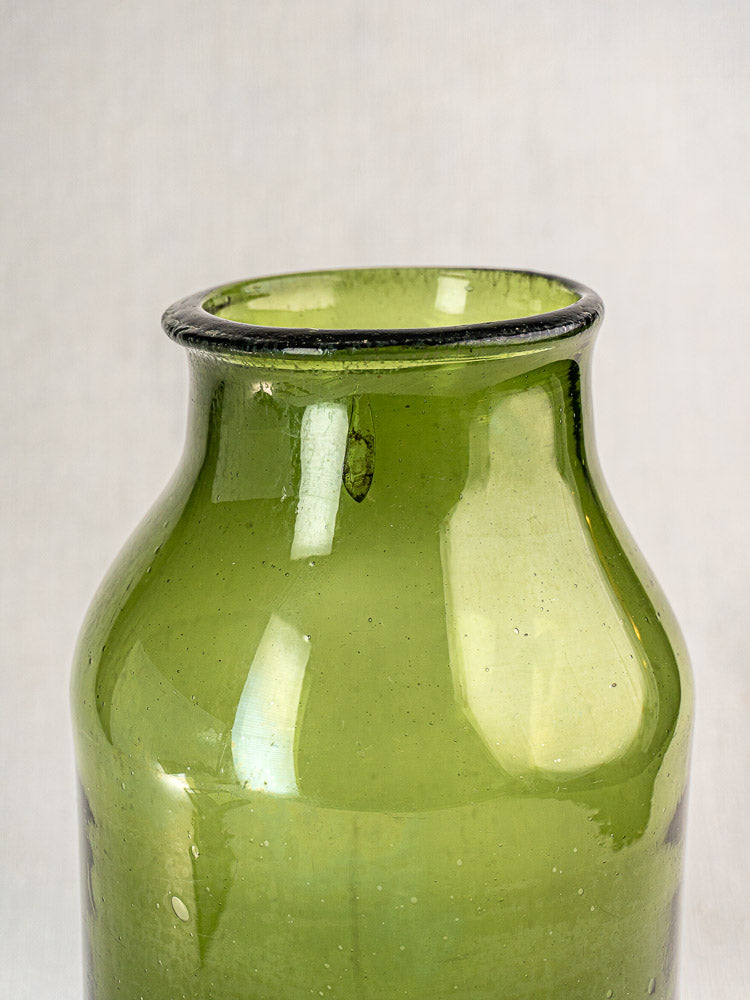 Antique French cornichon jar in hand-blown green glass, narrow neck and wide body, glossy with bubbles, on light gray background.