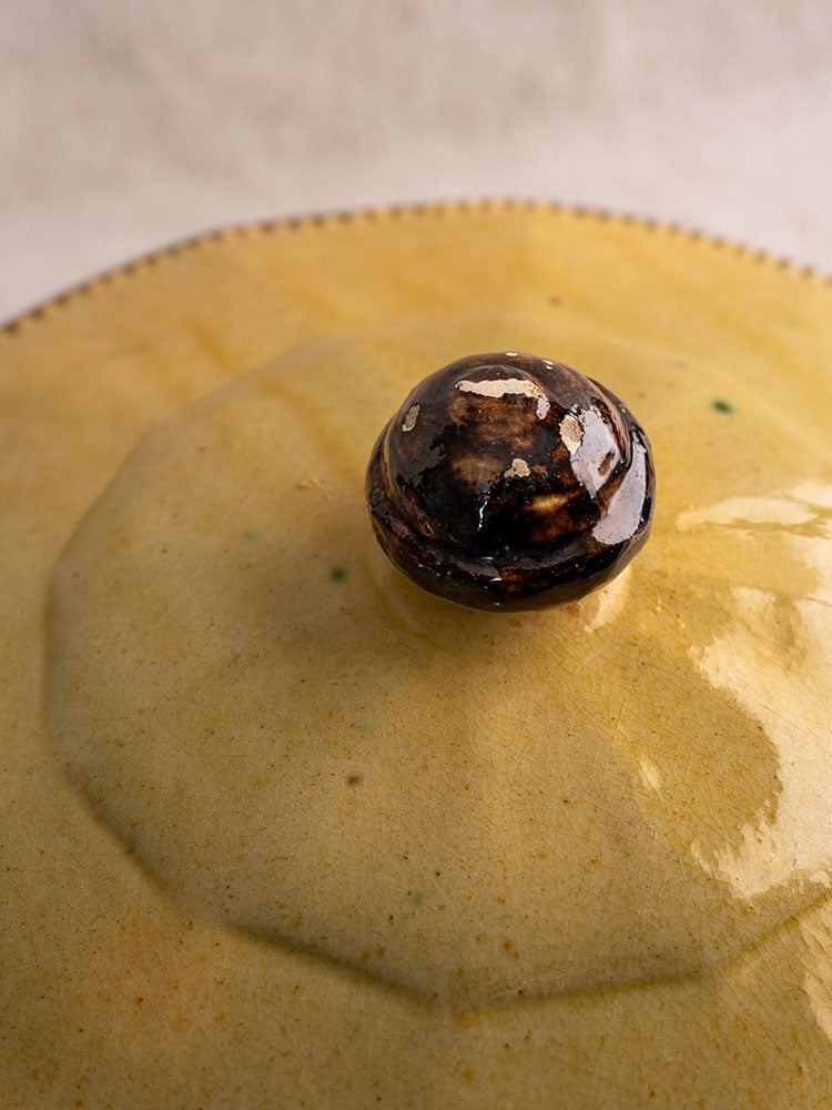 Mustard-glazed Vallauris pottery lid with green specks and dark brown knob, glossy finish, soft background.