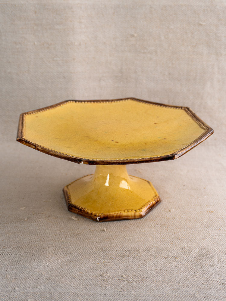 Mustard-glazed octagonal ceramic pedestal dish with brown edges, Vallauris Beretta et Cie, on neutral fabric backdrop.