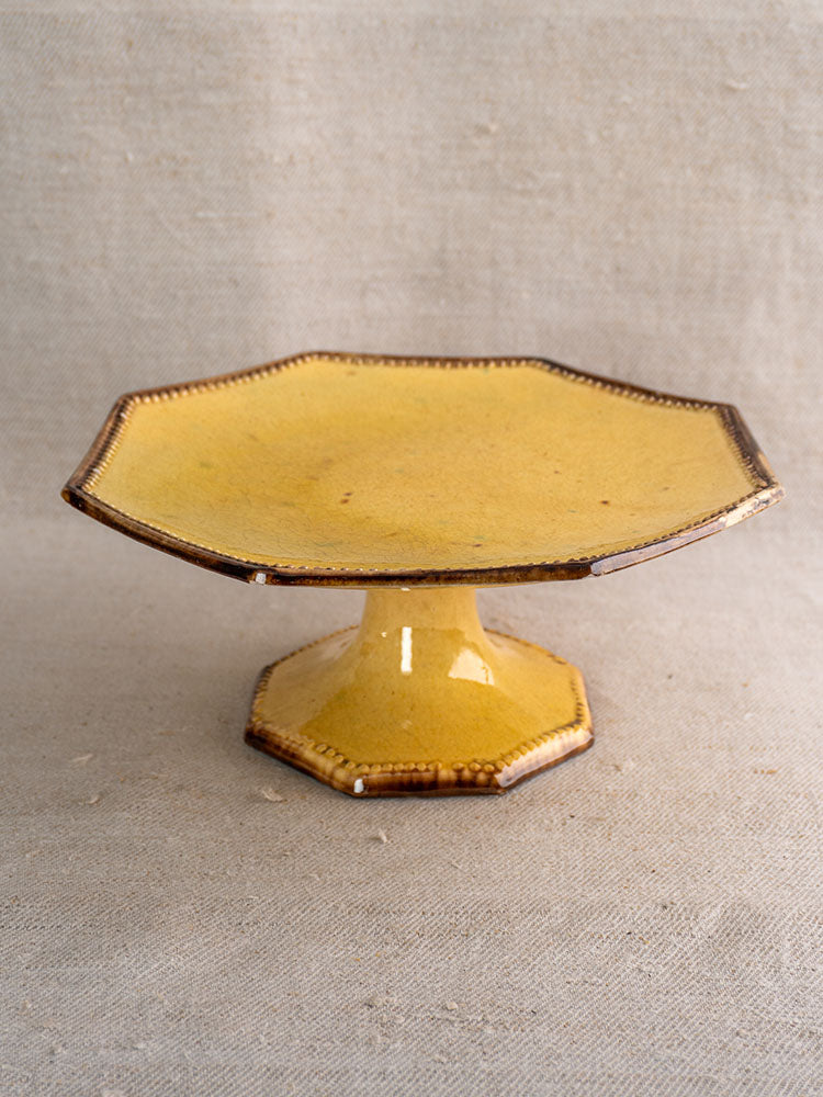 Mustard-glazed octagonal Vallauris ceramic cake stand with brown-edged rim, from Beretta et Cie set, on neutral fabric.