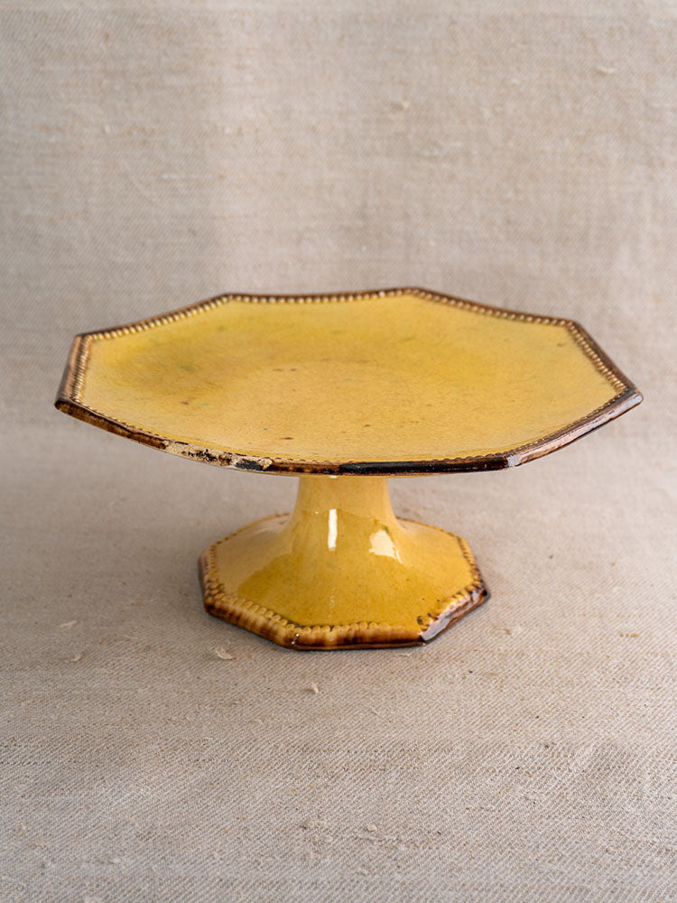 Mustard-glazed Vallauris Beretta et Cie cake stand with pedestal base and worn edges on beige fabric.