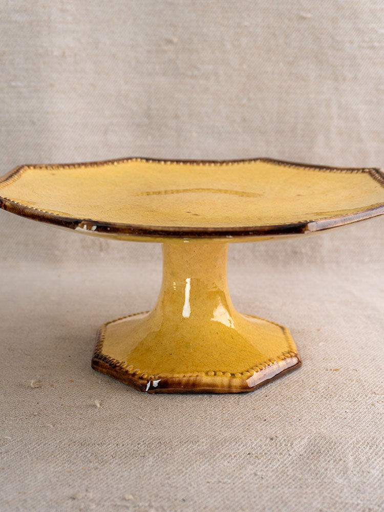Mustard glazed ceramic octagonal cake stand by Vallauris Beretta et Cie with brown rim on fabric background.