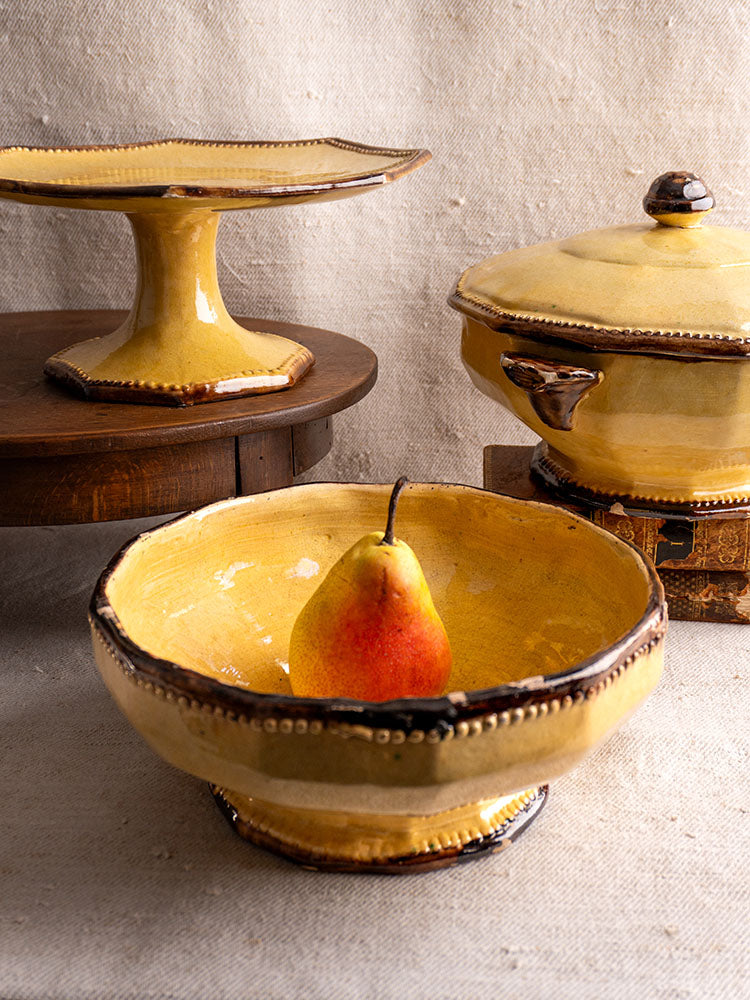Mustard-glazed Vallauris Beretta et Cie bowl with matching cake stand and lidded dish on neutral textured fabric.