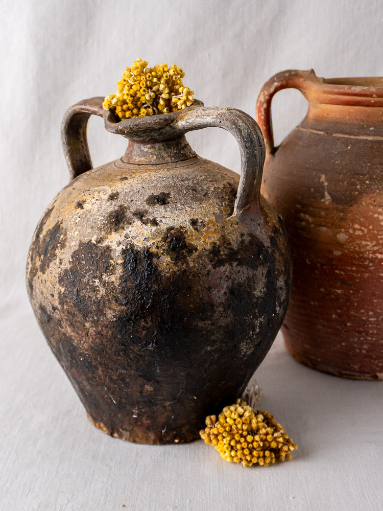 Antique French terracotta two-handled orjols with dried yellow flowers on a neutral fabric background.