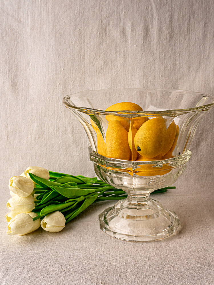 Antique French glass Coupe Médicis with yellow mangoes, pale fabric surface, and white tulips.