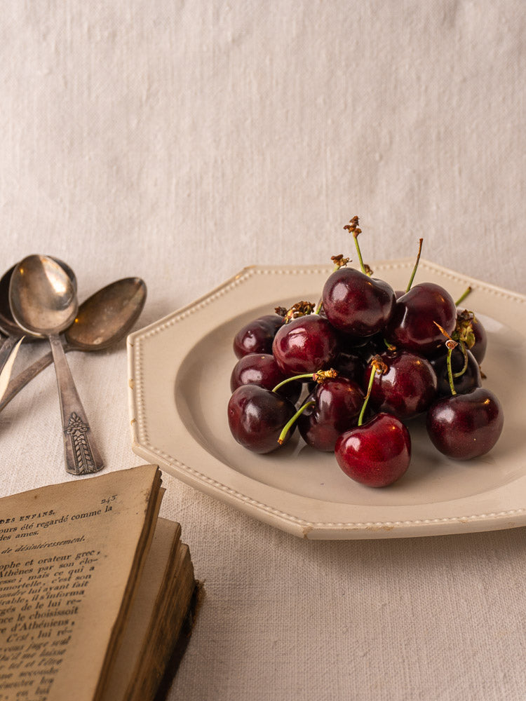 Antique French creamware plate with octagonal beaded rim, fresh cherries, beige cloth, vintage spoons, open antique book.