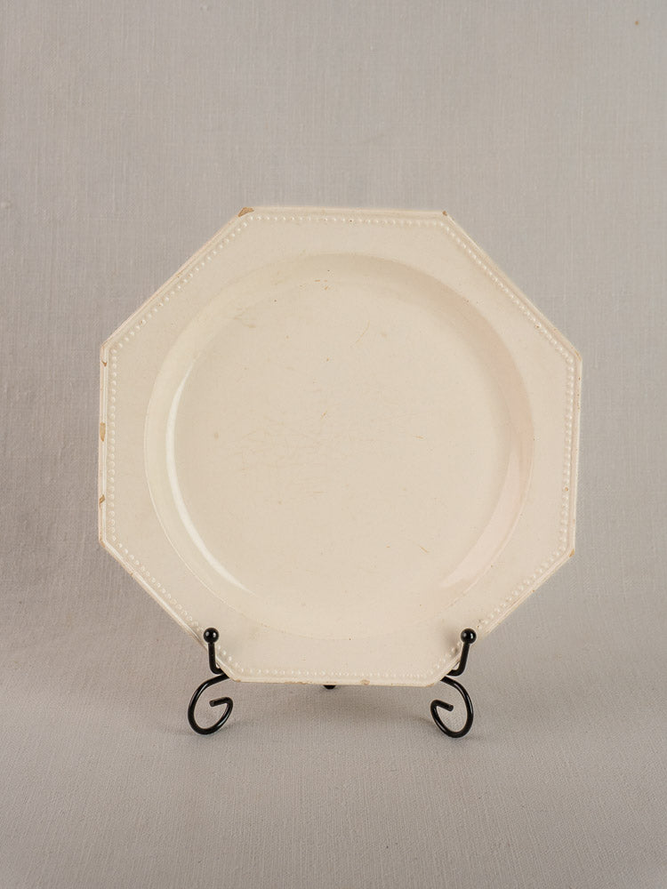 19th-century French creamware octagonal plate with beaded rim on black metal stand, neutral gray backdrop.