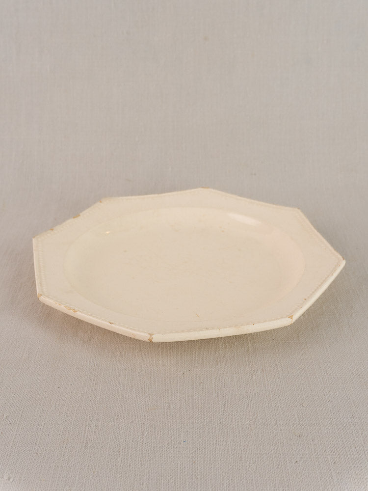 19th-century French creamware plate with octagonal beaded rim in off-white on light gray textured surface.