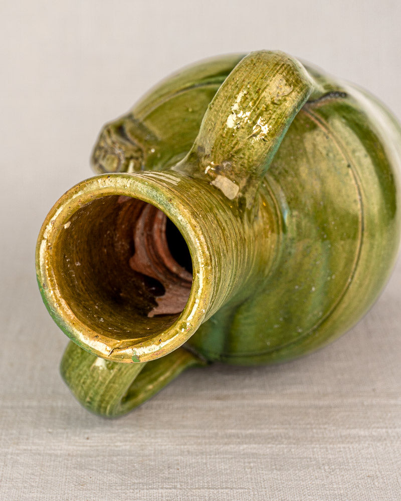 Green glazed antique French cruche with devotional medallion, dual handles, and wide mouth on neutral fabric background.