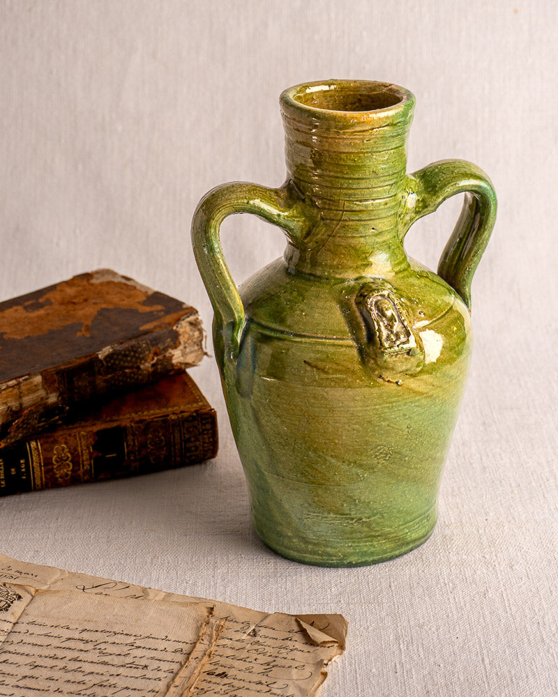 Antique French green glazed earthenware cruche with two handles and medallion beside vintage books and aged letter.