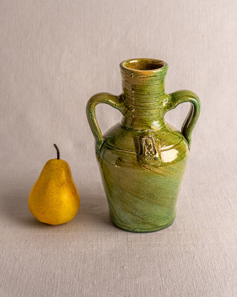 Antique French green glazed earthenware cruche with two handles and medallion, beside a yellow pear on neutral background.