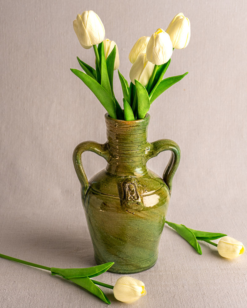 Green glazed French earthenware cruche with two handles holding white tulips, on a light grey background.