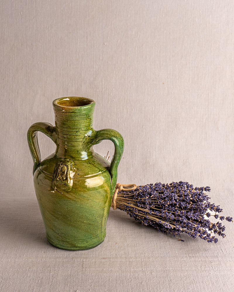 Antique French green glazed earthenware cruche with devotional medallion beside dried lavender on neutral fabric.