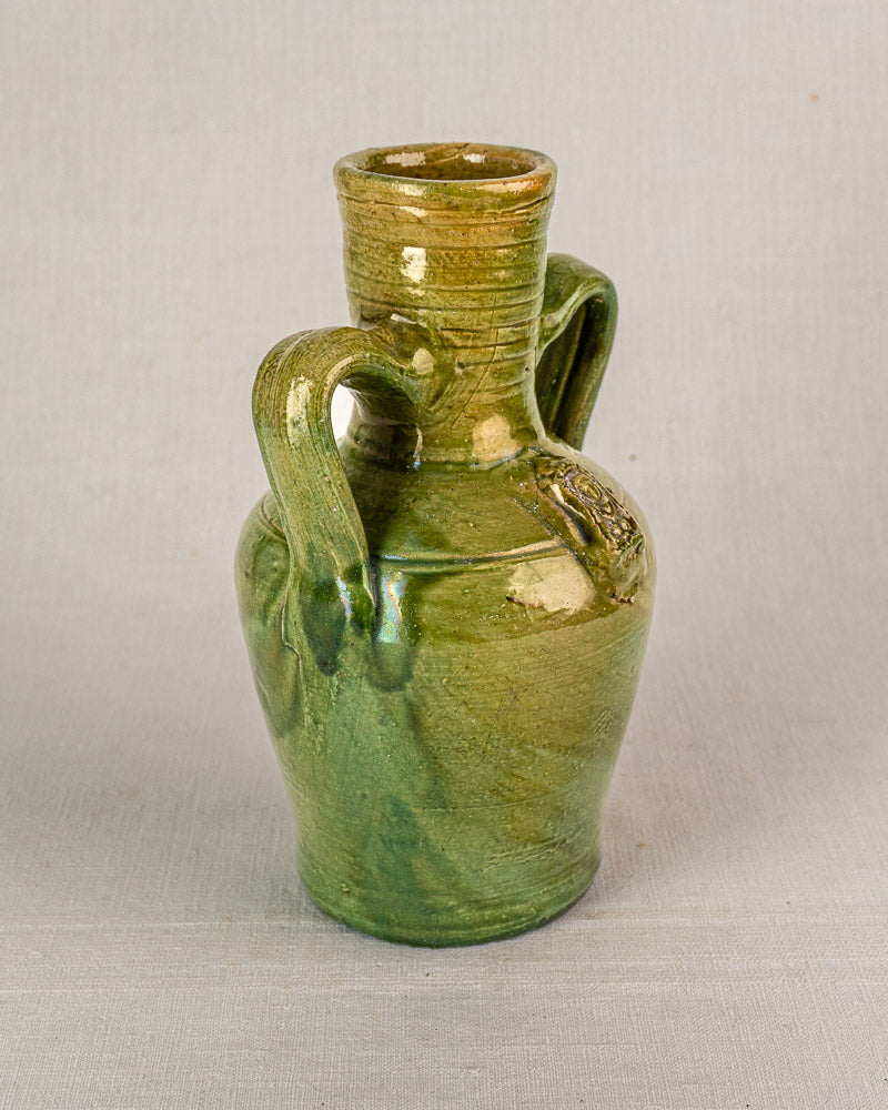 Antique French cruche in green glazed earthenware with two handles, narrow neck, and medallion on neutral background.