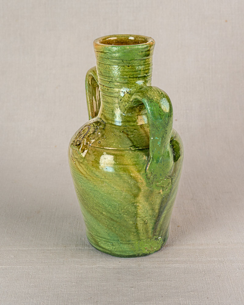 Antique French green-glazed earthenware cruche with two handles, narrow neck, textured shine; light background.