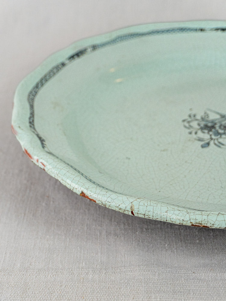 Antique French faience charger with celadon glaze, floral motif, visible cracks, and chipped rim on fabric background.