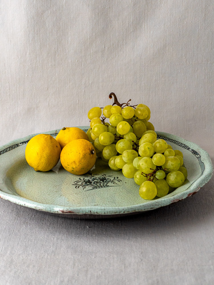 Antique Cul Noir French faience charger in celadon with lemons and green grapes on beige fabric.
