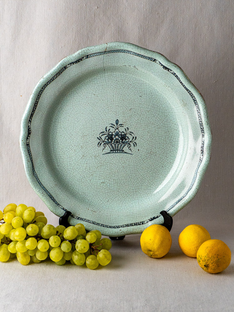 Antique French faience charger with celadon glaze and black floral motif on display stand, lemons and grapes in front.