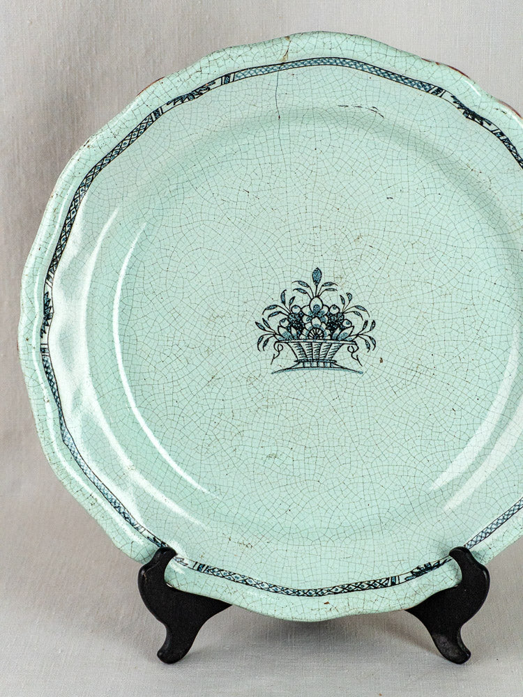 Pale blue celadon French faience charger with crackled glaze, scalloped edge, and black floral basket motif.
