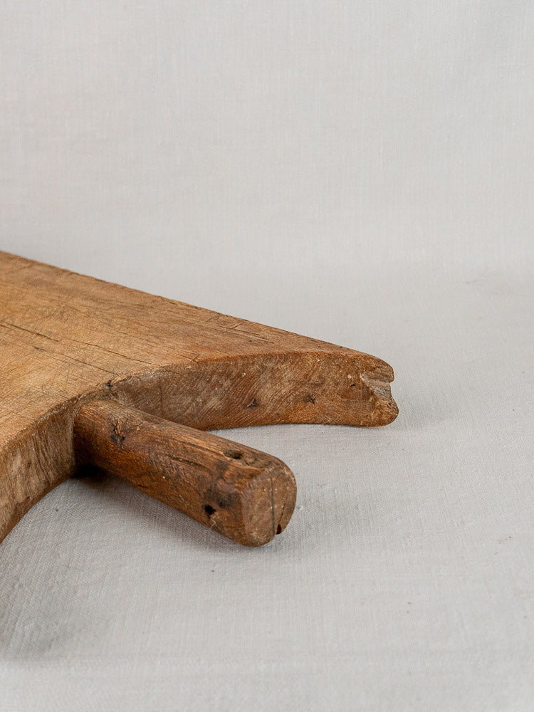 Antique French hand-carved wood cutting board with wing-shouldered edge and rustic handle on light fabric.