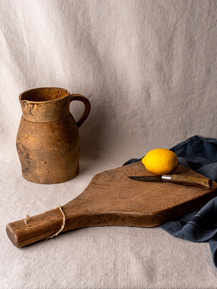 Antique French hand-carved chestnut cutting board with tapered handle, upright, highlighting wood grain and knife marks.