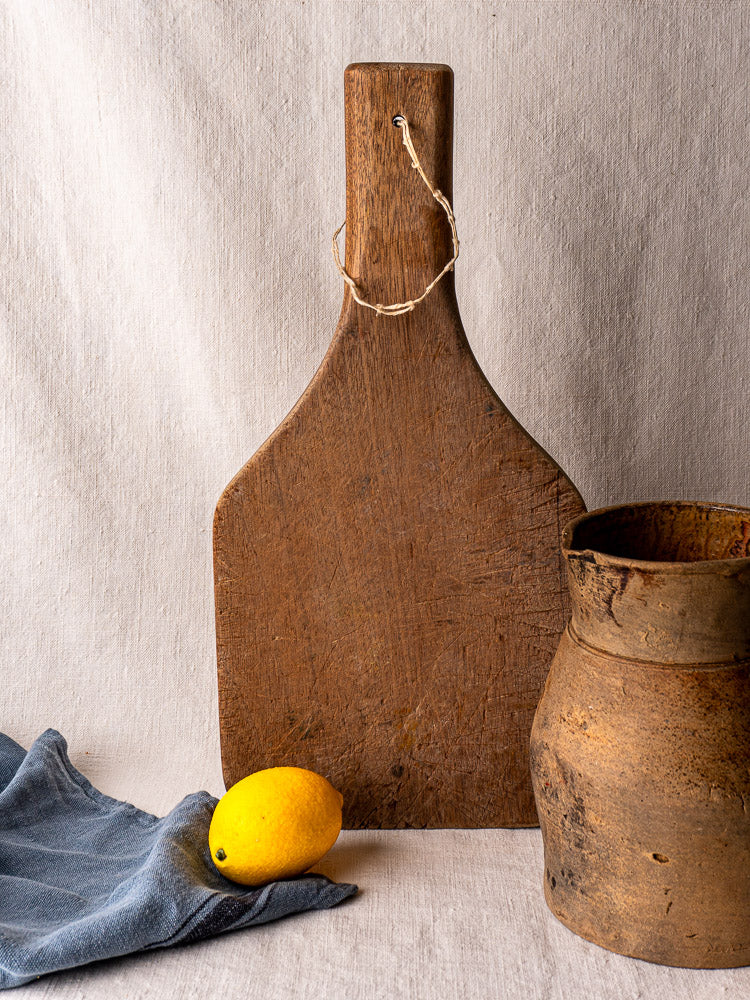 Antique French chestnut cutting board with tapered handle, shown upright beside a lemon and weathered clay pot.