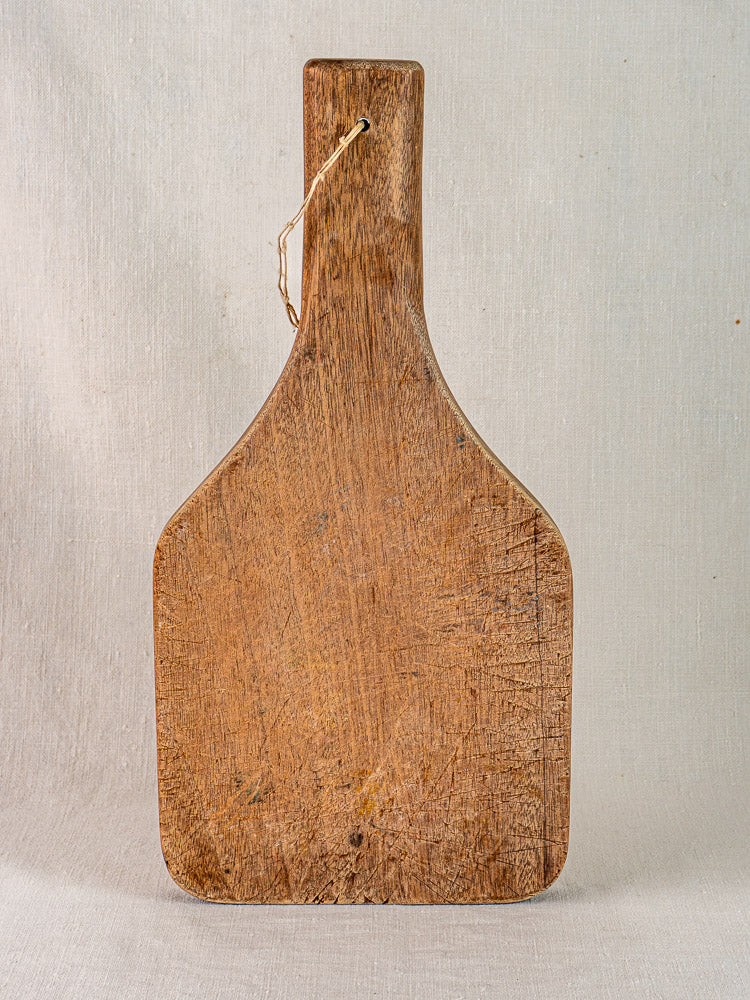 Antique French hand-carved chestnut cutting board with tapered handle, upright, highlighting wood grain and knife marks.