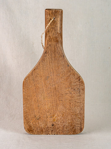 Antique French hand-carved chestnut cutting board with tapered handle, upright, highlighting wood grain and knife marks.