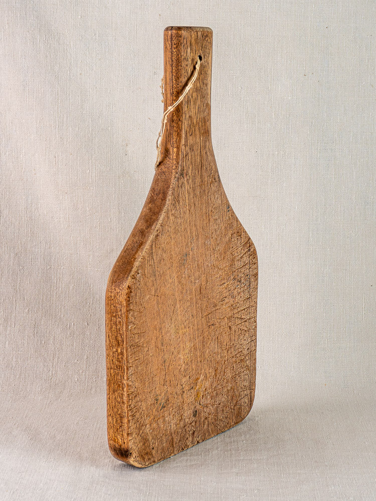Antique French chestnut cutting board with tapered handle, upright against a neutral fabric backdrop.