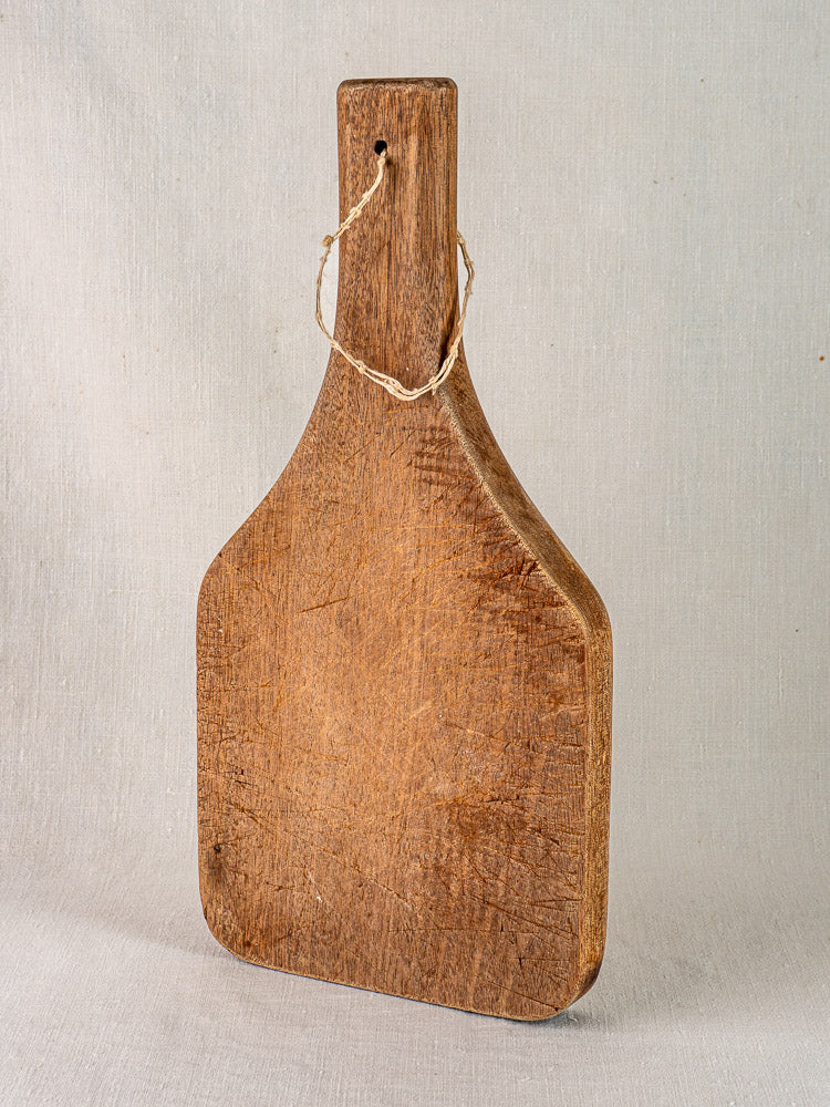 Antique French chestnut cutting board with tapered handle, upright against a neutral fabric backdrop, rustic Provence style.