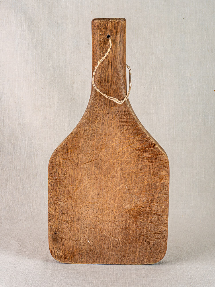 Antique French chestnut cutting board with tapered handle, hand carved and showing rustic texture against a light fabric backdrop.
