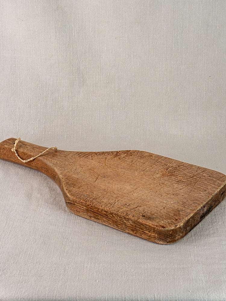Antique French chestnut cutting board with a tapered handle displayed on light fabric.