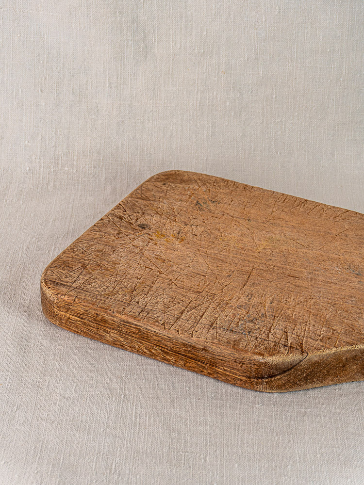 Antique French chestnut cutting board with tapered handle, hand carved and rustic, on a light textured surface.