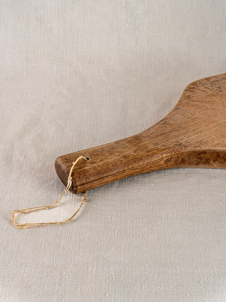 Antique French chestnut cutting board with tapered handle and twine loop, displayed on light beige fabric.