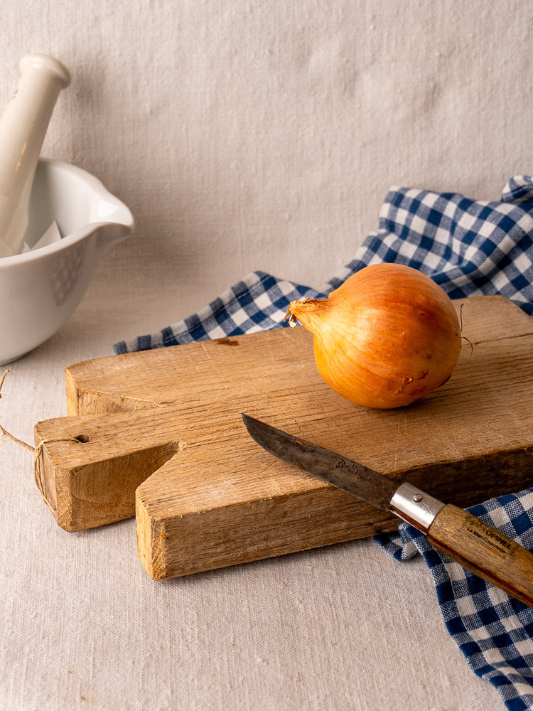 Hand-carved antique French oak cutting board with whole onion and knife, blue checkered cloth, white mortar and pestle behind.