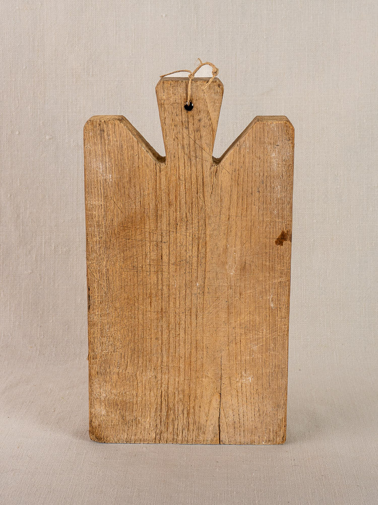 Antique French oak cutting board with notched top and hanging hole, upright on a neutral fabric backdrop.