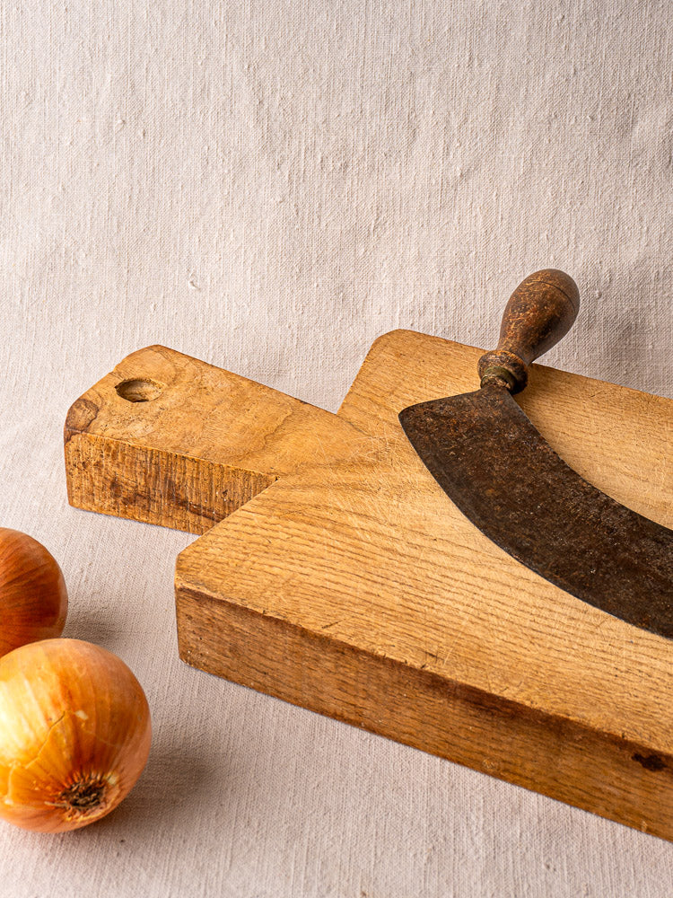 Antique French wooden cutting board with vintage curved chopping knife and two brown onions on beige cloth.