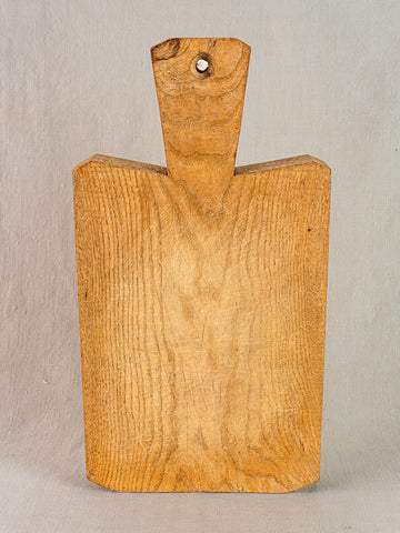 Antique French cutting board, hand-carved thick wood with short wide handle and top hole, upright on neutral background.