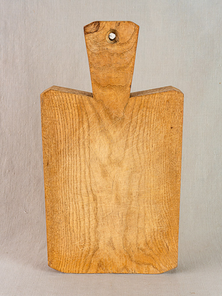 Antique French cutting board, hand-carved thick wood with short wide handle and top hole, upright on neutral background.