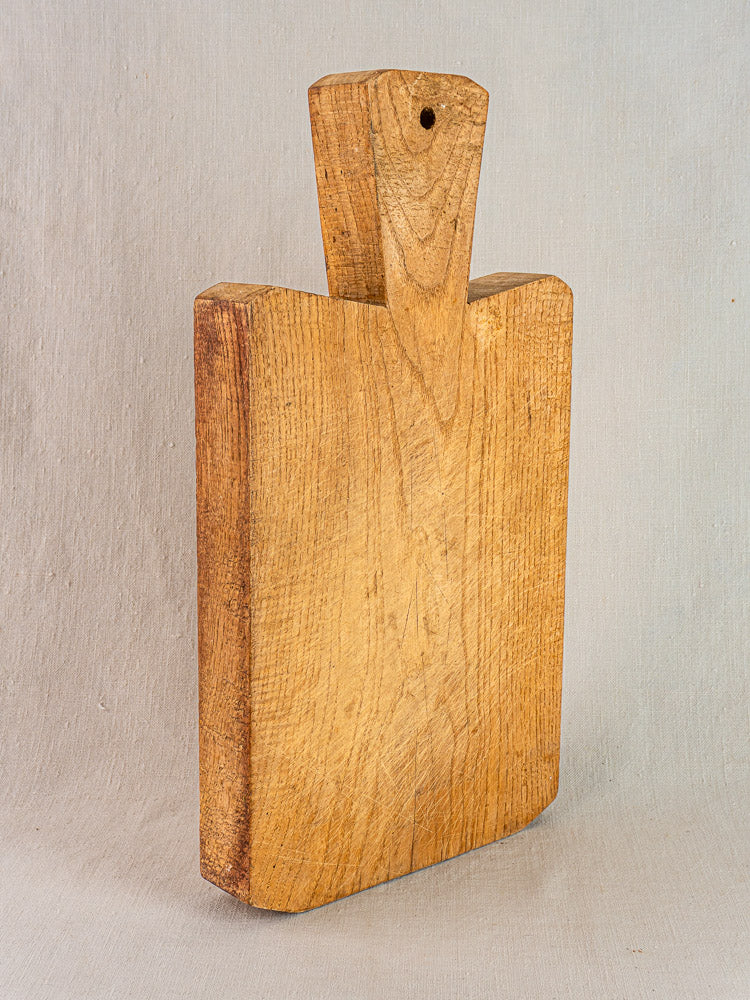 Antique French hand-carved wood cutting board with visible grain, short handle, and hanging hole on light textured surface.