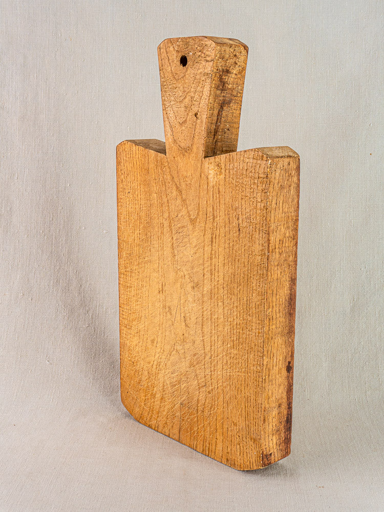 Antique French hand-carved wooden cutting board with angled handle, displayed upright on neutral fabric background.