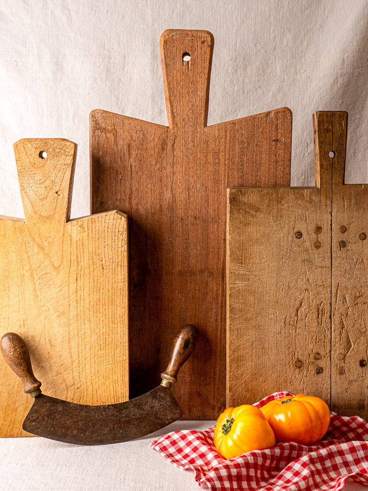 Antique French hardwood cutting board with rustic boards, vintage chopper, and yellow tomatoes on a checkered cloth.