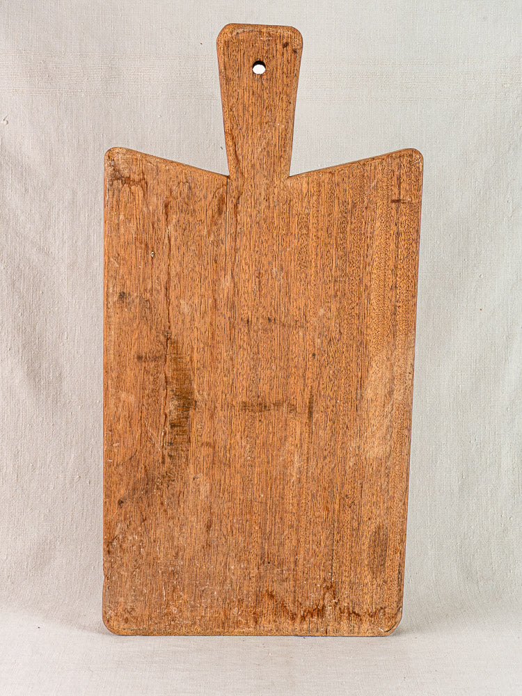 Antique French hardwood cutting board with handle and top hole, upright on a light textured background.