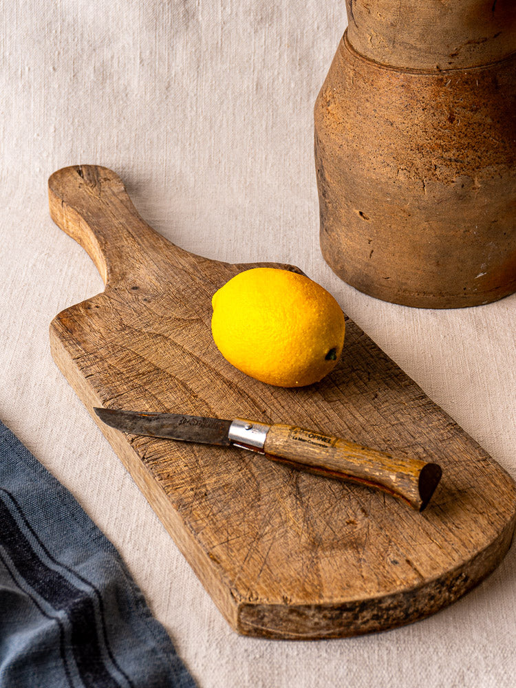 Antique French oak or chestnut cutting board with lemon, rustic knife, brown clay jug, and blue-striped cloth on beige fabric.