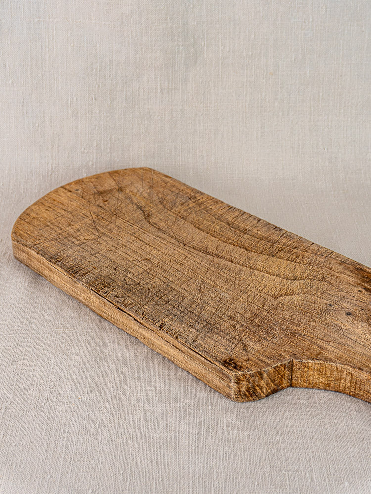 Antique French oak or chestnut cutting board with rounded end, visible grain and knife marks on beige linen fabric.